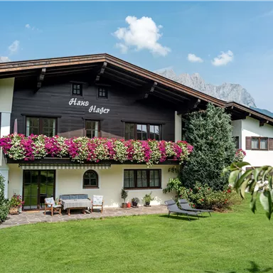 A charming house with a blooming balcony and a well-maintained garden. In the background, mountains and a clear sky can be seen.