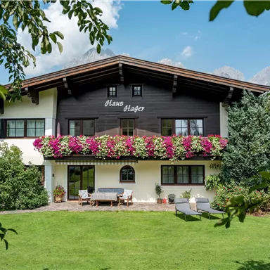 A charming house with a flower balcony and a well-maintained garden. In the background, green trees and a blue sky can be seen.