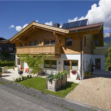 A beautiful wooden house with a landscaped garden and a stone driveway. In the background, mountains and a clear sky can be seen.