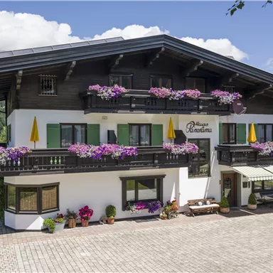 A charming alpine house with blooming balcony plants. The facade is white, with colorful accents and a well-maintained outdoor area.