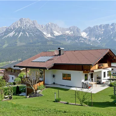 Eine malerische Landschaft mit einem modernen Haus und den Bergen im Hintergrund. Der Garten ist grün und einladend, ideal für eine ruhige Auszeit.