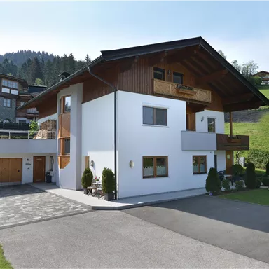 Ein modernes Haus mit Holzelementen und breiter Auffahrt. Die Umgebung ist grün mit Hügeln im Hintergrund.