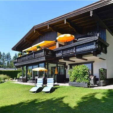 A modern house with a large garden and sun loungers. Orange umbrellas are on the terrace and it is surrounded by trees.