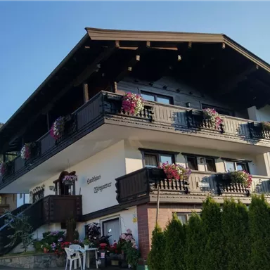 A picturesque house in alpine style with flowering balcony boxes. Green mountains are visible in the background.