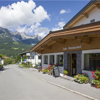 Ein charmantes Restaurant mit Holzfassade befindet sich an einer ruhigen Straße. Im Hintergrund sind beeindruckende Berge und ein blauer Himmel zu sehen.