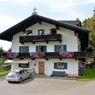 Ein charmantes Alpenhaus mit blühenden Balkonen. Vor dem Haus steht ein Auto, umgeben von einer grünen Landschaft.