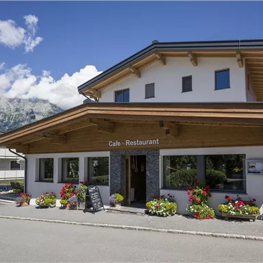 Ein gemütliches Café-Restaurant mit Holzverkleidung und bunten Blumenbeeten. Im Hintergrund sind die Berge und der blaue Himmel zu sehen.