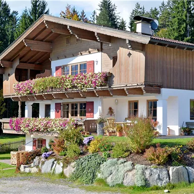 Ein typisches Alpenhaus mit Holzverkleidung und bunten Blumen auf dem Balkon. Umgeben von grünen Wiesen und Bäumen.