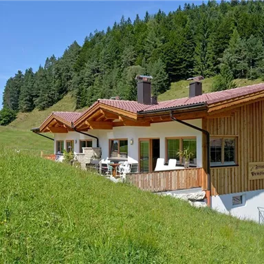 Ein gemütliches Haus im alpinen Stil, umgeben von grünen Wiesen und Bäumen. Im Hintergrund erstreckt sich eine malerische Berglandschaft.