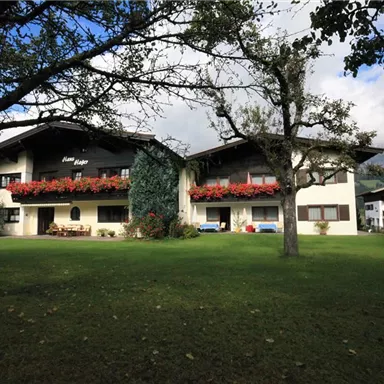 A charming house with flowering balconies in a green surroundings. The sky is partly cloudy and the landscape is idyllic.