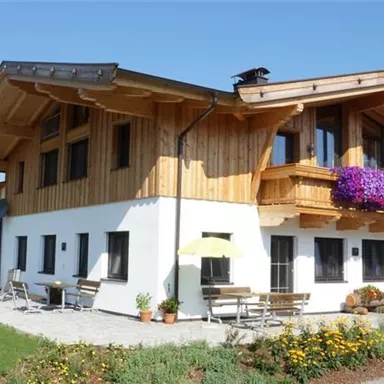 A modern wooden house with a large garden and colorful flowers. In the background, green hills and a blue sky can be seen.