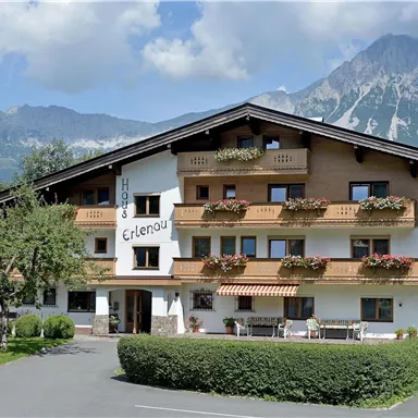 A charming hotel with blooming balconies and a beautiful mountain landscape in the background. The sky is clear, and the surroundings are green and inviting.