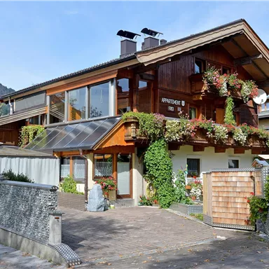 A charming wooden house in the Alps with blooming flowers in front of the window. In the background, mountains can be seen and the sky is clear.