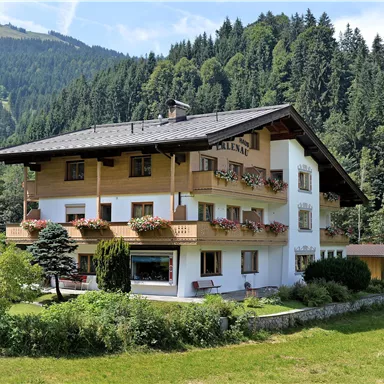 A charming house with a wooden balcony amidst green meadows and trees. In the background, mountains can be seen, creating a picturesque atmosphere.