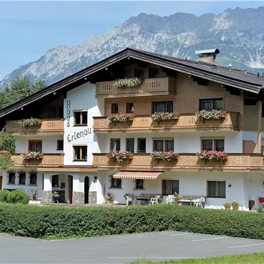 A charming hotel with a wooden facade and blooming balconies. In the background, picturesque mountains and a clear sky can be seen.
