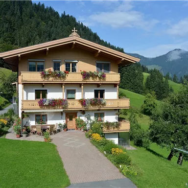 Ein schönes Gebäude im alpinen Stil, umgeben von grünen Wiesen und Bergen. Das Haus hat Balkone mit Blumen und einen gepflegten Garten.