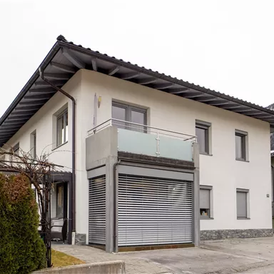 Ein modernes Wohnhaus mit weißer Fassade und einem Balkon. Die Umgebung ist ruhig und wird von Bergen umgeben.