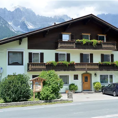 Ein traditionelles Haus mit einem großen Balkon und vielen Blumen. Im Hintergrund sind Berge und eine grüne Landschaft zu sehen.