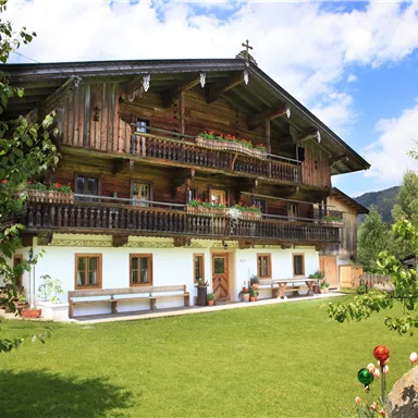 Ein traditionelles Holzhaus mit mehreren Balkonen in einer grünen Umgebung. Der Himmel ist blau mit einigen Wolken und die Landschaft ist ruhig.
