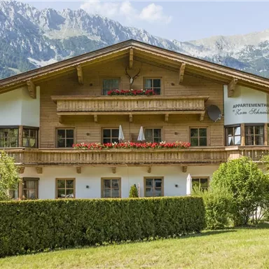 Ein schönes Holzhaus mit Balkonen und blühenden Blumenkästen. Die Umgebung ist von grünen Wiesen und Bergen umgeben.