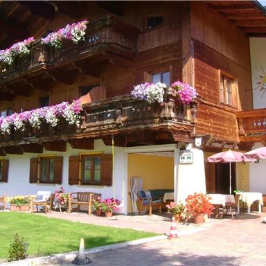 A picturesque wooden house with blooming balconies. In the foreground, a well-maintained garden and sunny seating areas can be seen.