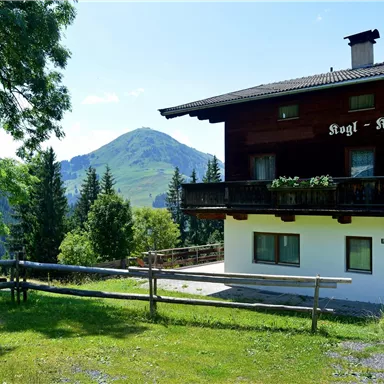 Ein traditionelles Chalet mit einem großen Balkon. Dahinter erhebt sich ein grüner Berg unter klarem blauem Himmel.