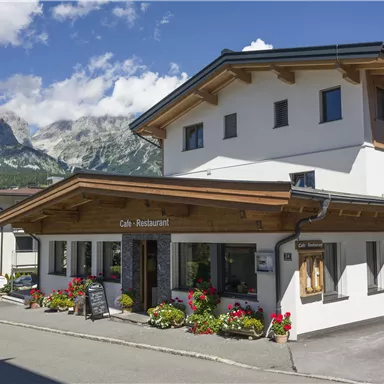 Ein einladendes Restaurant mit einer gemütlichen Fassade und blühenden Pflanzen. Im Hintergrund sind majestätische Berge und ein blauer Himmel zu sehen.