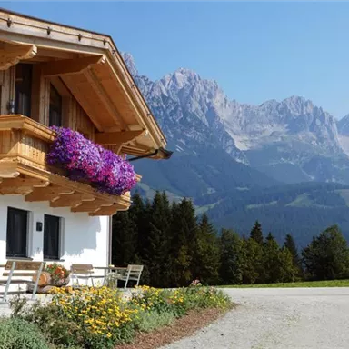 A charming house with a wooden balcony and colorful flowers. In the background, impressive mountains and a clear sky can be seen.