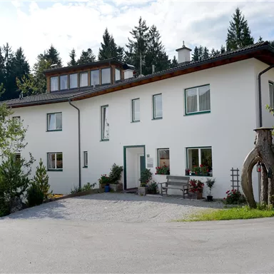 A modern, white house with large windows and an elegant entrance. Surrounded by trees and a paved path.