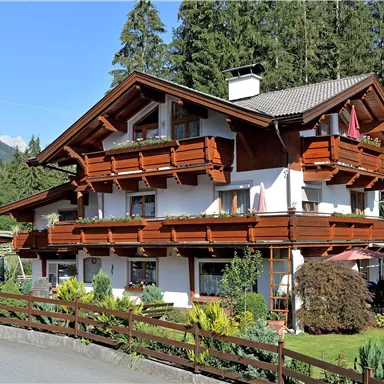 A charming wooden house in alpine style, surrounded by trees and beautiful vegetation. The balconies are decorated with flowers, and the building radiates an inviting atmosphere.