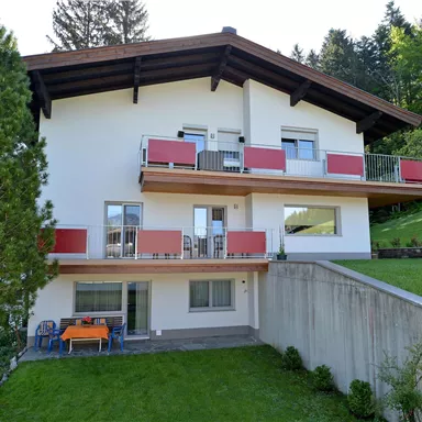 Ein modernes Haus mit mehreren Balkonen und roten Geländern. Es ist von grünen Wiesen und Bäumen umgeben.