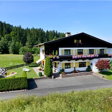 Ein gemütliches Haus mit bunten Blumen und umgeben von einer grünen Wiese. Im Hintergrund sind hohe Bäume und Berge zu sehen.