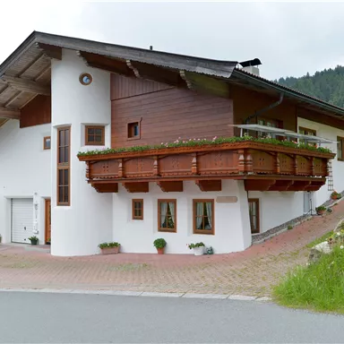 Ein modernes Haus mit einem Balkon aus Holz und einem stilvollen Design. Es steht in einer grünen Umgebung mit sanften Hügeln im Hintergrund.