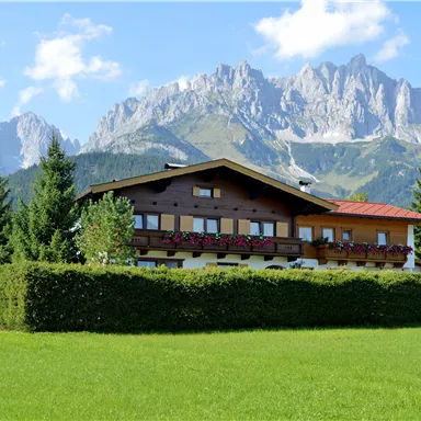 Ein traditionelles chaletartiges Haus umgeben von grünen Wiesen und Bäumen. Im Hintergrund erheben sich majestätische Berge unter einem blauen Himmel.