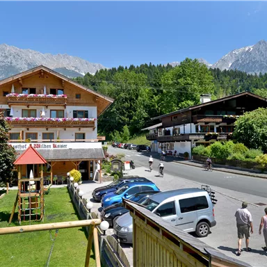 A picturesque building with a balcony and beautiful garden, surrounded by mountains. People are strolling along the street under a blue sky.