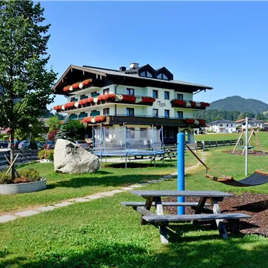 Ein gemütliches Gebäude mit Balkonen und bunten Blumen. Im Vordergrund gibt es eine Spielwiese mit einer Schaukel und einer Sitzecke.