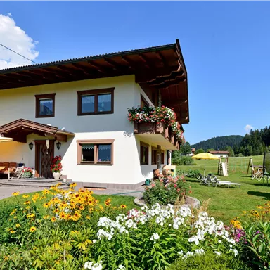 Ein charmantes Haus mit Blumen im Vordergrund und einer schönen Berglandschaft im Hintergrund. Der Himmel ist klar und die Umgebung ist grün und einladend.