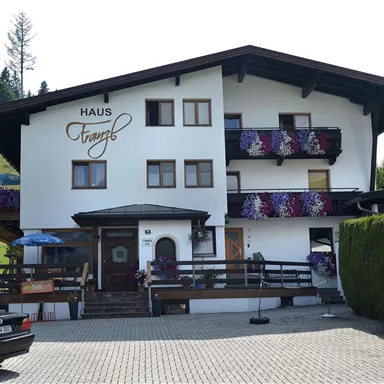 A cozy house with blooming balconies and a clear blue sky. In front of the house, several cars are parked on a paved parking lot.
