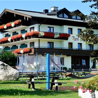 Ein modernes Gebäude mit Balkonblumen und einem freundlichen Garten. Im Vordergrund befinden sich Sitzgelegenheiten und eine Hängematte.