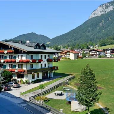 Ein malerisches Landhaus mit Blumenkästen steht in einer grünen Landschaft. Im Hintergrund sind Berge und eine ruhige Nachbarschaft zu sehen.