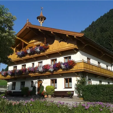Ein traditionelles Holzhaus mit einem schönen Balkon, der mit bunten Blumen geschmückt ist. Umgeben von grünen Bäumen und Bergen im Hintergrund.