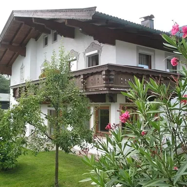 Ein idyllisches Haus im alpinen Stil mit einem Balkon aus Holz. Der Garten ist grün und blühend, umgeben von Bäumen und Pflanzen.