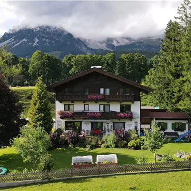 Ein gemütliches Haus umgeben von Grün und Bäumen, mit Bergen im Hintergrund. Der Garten ist einladend und ideal zum Entspannen.