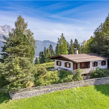 Ein charmantes Haus in den Bergen, umgeben von üppigem Grün und hohen Bäumen. Im Hintergrund sind die majestätischen Berge zu sehen und der Himmel ist klar und blau.