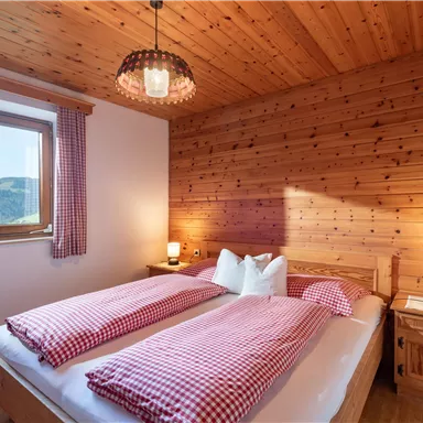 Ein gemütliches Schlafzimmer aus Holz mit einem Fenster, das einen Blick auf die Berge bietet. Die zwei Betten sind mit karierten Decken und Kissen dekoriert.