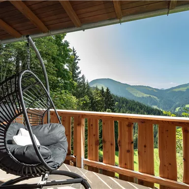 Ein schaukelnder Stuhl auf einer Terrasse mit Blick auf grüne Berge. Der Himmel ist klar und sonnig, ideal zum Entspannen.