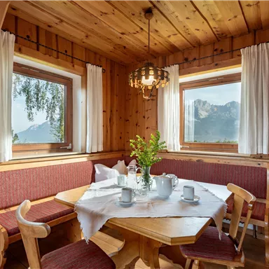 Ein gemütlicher Essbereich aus Holz mit einem runden Tisch und Sitzbänken. Durch die Fenster sieht man die Berge und das klare Wetter.