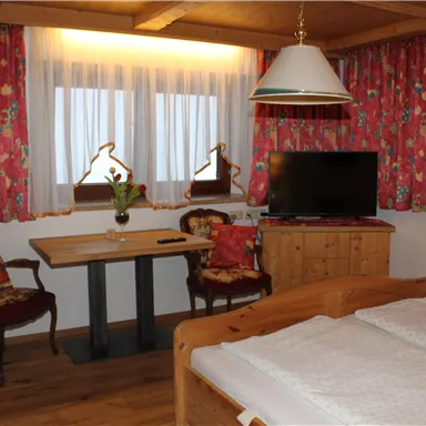 Ein gemütliches Zimmer mit Holzdetails, einem Doppelbett und einem Tisch mit zwei Stühlen. Vor den Fenstern hängen farbenfrohe Vorhänge, und ein Fernseher steht an der Wand.