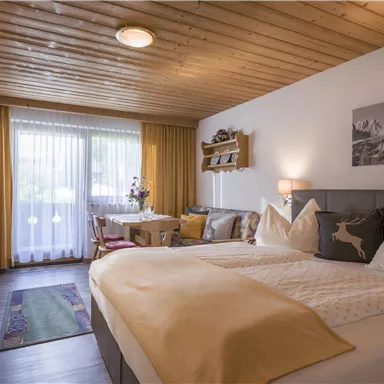 Ein gemütliches Hotelzimmer mit einem großen Doppelbett und Holzdecke. Es gibt Fenster mit Vorhängen und einen kleinen Essbereich.