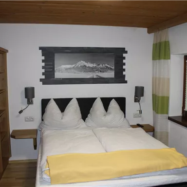 Ein gemütliches Schlafzimmer mit einem Doppelbett, Holzschränken und moderner Dekoration. Die Wände sind hell, und die Fenster haben farbige Vorhänge.